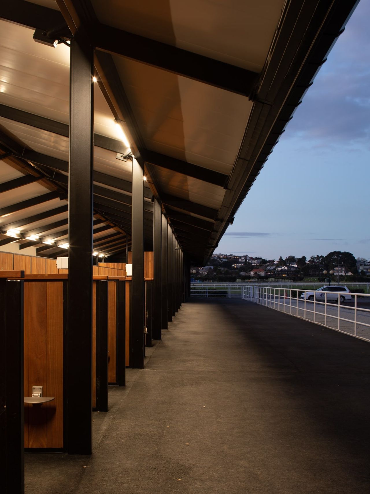 Covered walkway alongside horse stalls at Auckland Racing Club stables, lit by overhead lights during dusk