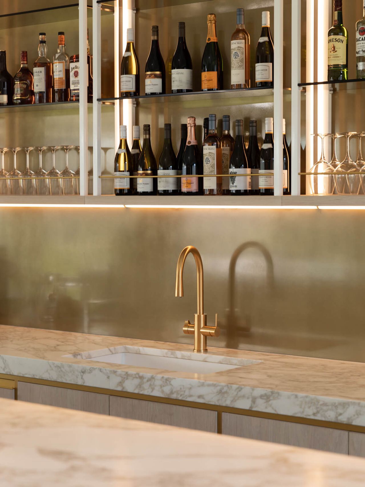 Elegant hospitality interior design at Cambridge Stud Farm, featuring a luxurious marble countertop with gold veining, a brushed gold faucet, and a backlit bar display with premium wine and spirits, highlighting sophisticated architectural and interior finishes.