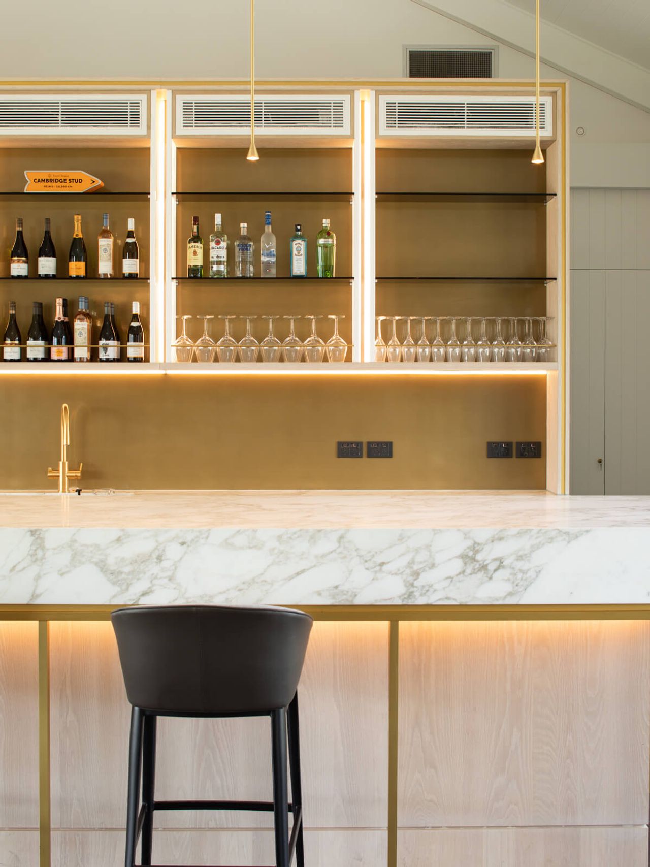 Modern hospitality bar design at Cambridge Stud Farm featuring a marble countertop with subtle veining, gold accents, backlit shelving for wine and spirits, and a sleek black bar stool, emphasizing luxury and contemporary architectural elements.