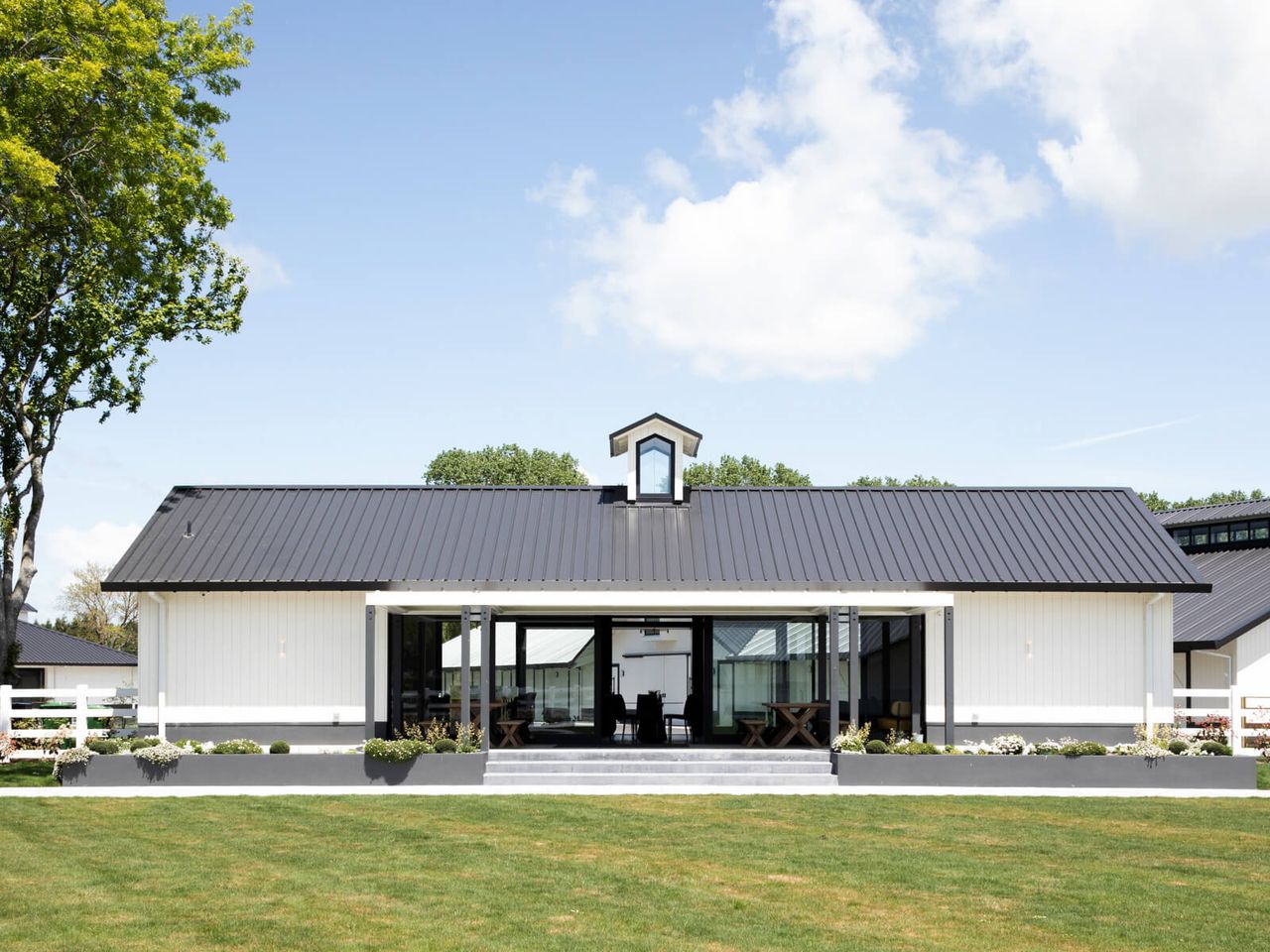 Front view of Cambridge Stud Farm's hospitality building, showcasing equine architecture with a black pitched roof, white board-and-batten cladding, large glass doors, and landscaped surroundings, blending rural charm with modern design in New Zealand.