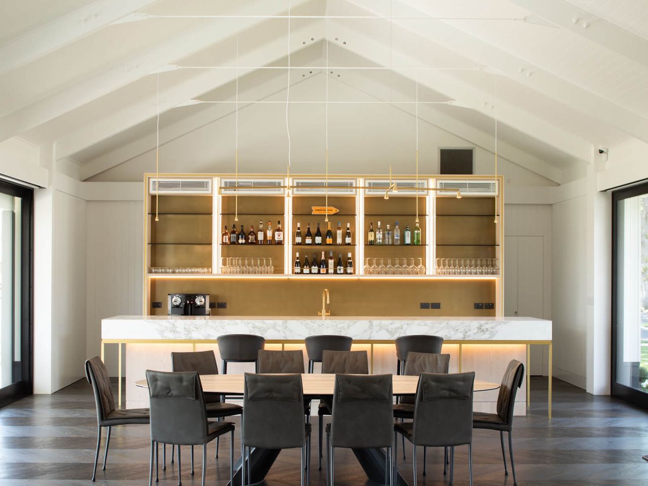 Luxurious interior of Cambridge Stud Farm's hospitality area, featuring a marble-topped bar with gold accents, backlit shelving, a contemporary dining table with black chairs, and vaulted ceilings, blending modern design with architectural elegance in New Zealand.