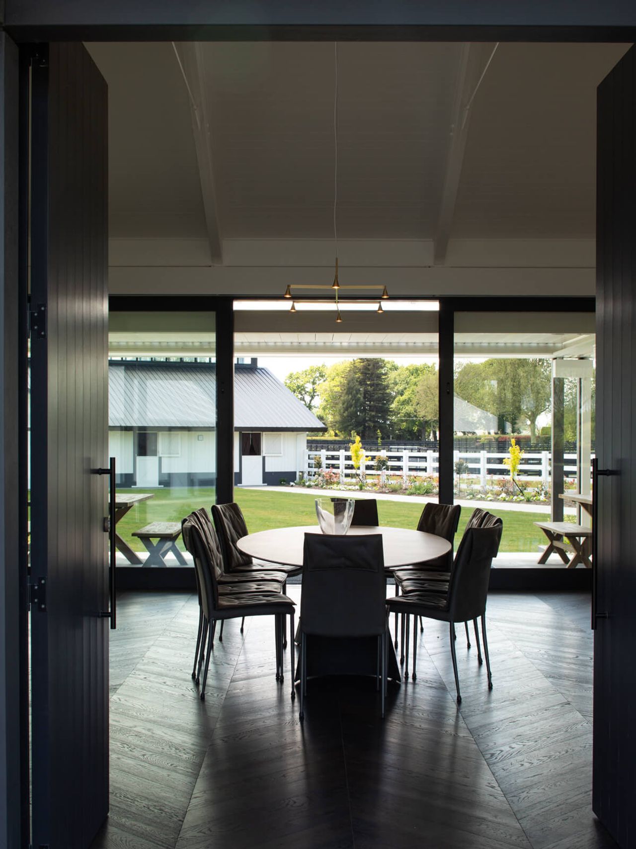 View of the dining area at Cambridge Stud Farm, featuring modern equine architecture with a round table, black chairs, and large glass doors opening to the landscaped outdoor space and surrounding stables, showcasing a seamless blend of functionality and elegant design in New Zealand.