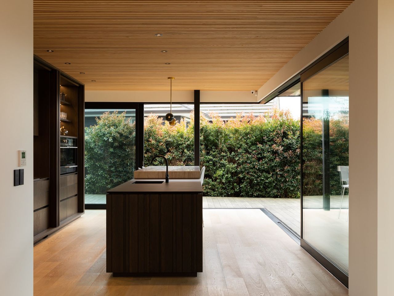 Open-plan kitchen in the Karaka Lake Showhome featuring natural wood finishes, floor-to-ceiling glass doors, and seamless indoor-outdoor flow. A stunning example of modern home design and interior architecture by Young+Richards+ in New Zealand.
