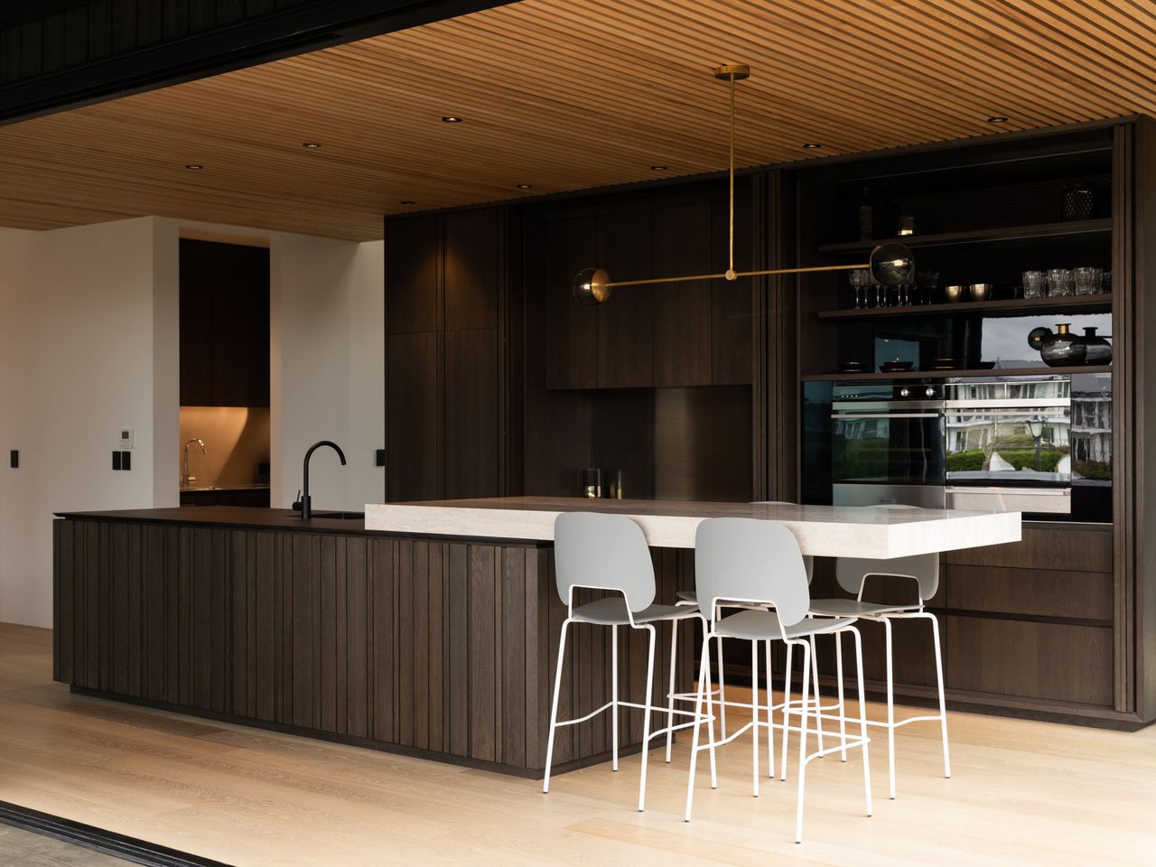 Modern kitchen interior of the Karaka Lake Showhome featuring dark wood cabinetry, sleek black fixtures, and a minimalist island with white bar stools. Designed by Young+Richards+, this space highlights premium interior architecture and home design in New Zealand.