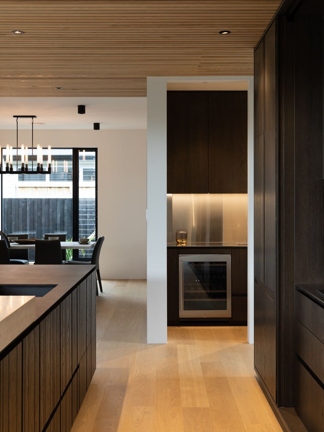 Open-plan kitchen and dining area in the Karaka Lake Showhome featuring dark wood cabinetry, integrated wine fridge, and elegant pendant lighting. Showcasing modern architectural design and sophisticated interiors by Young+Richards+ in New Zealand.