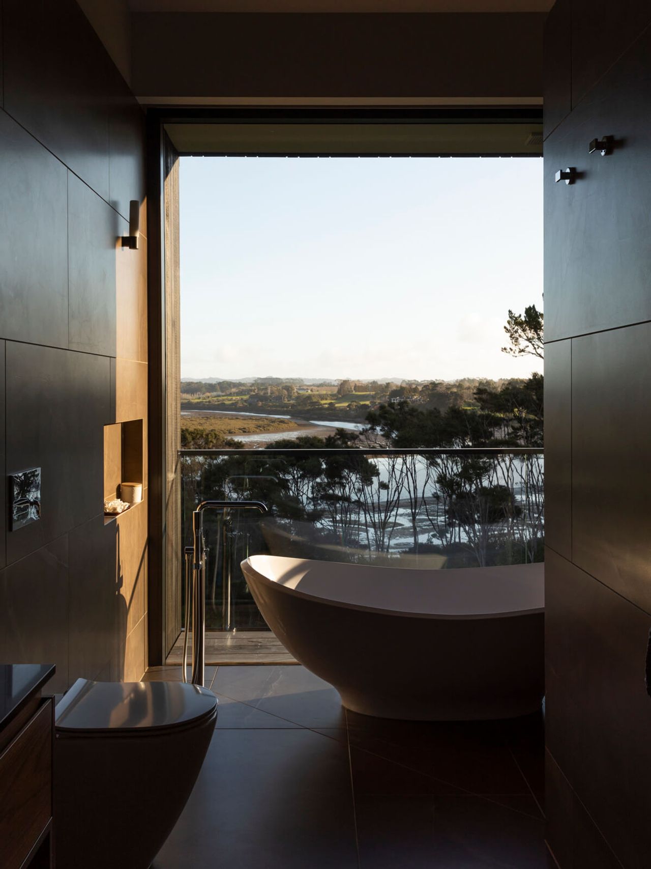 Luxurious modern bathroom featuring a freestanding bathtub with stunning floor-to-ceiling views of the New Zealand countryside and river, blending contemporary interior design with natural serenity.
