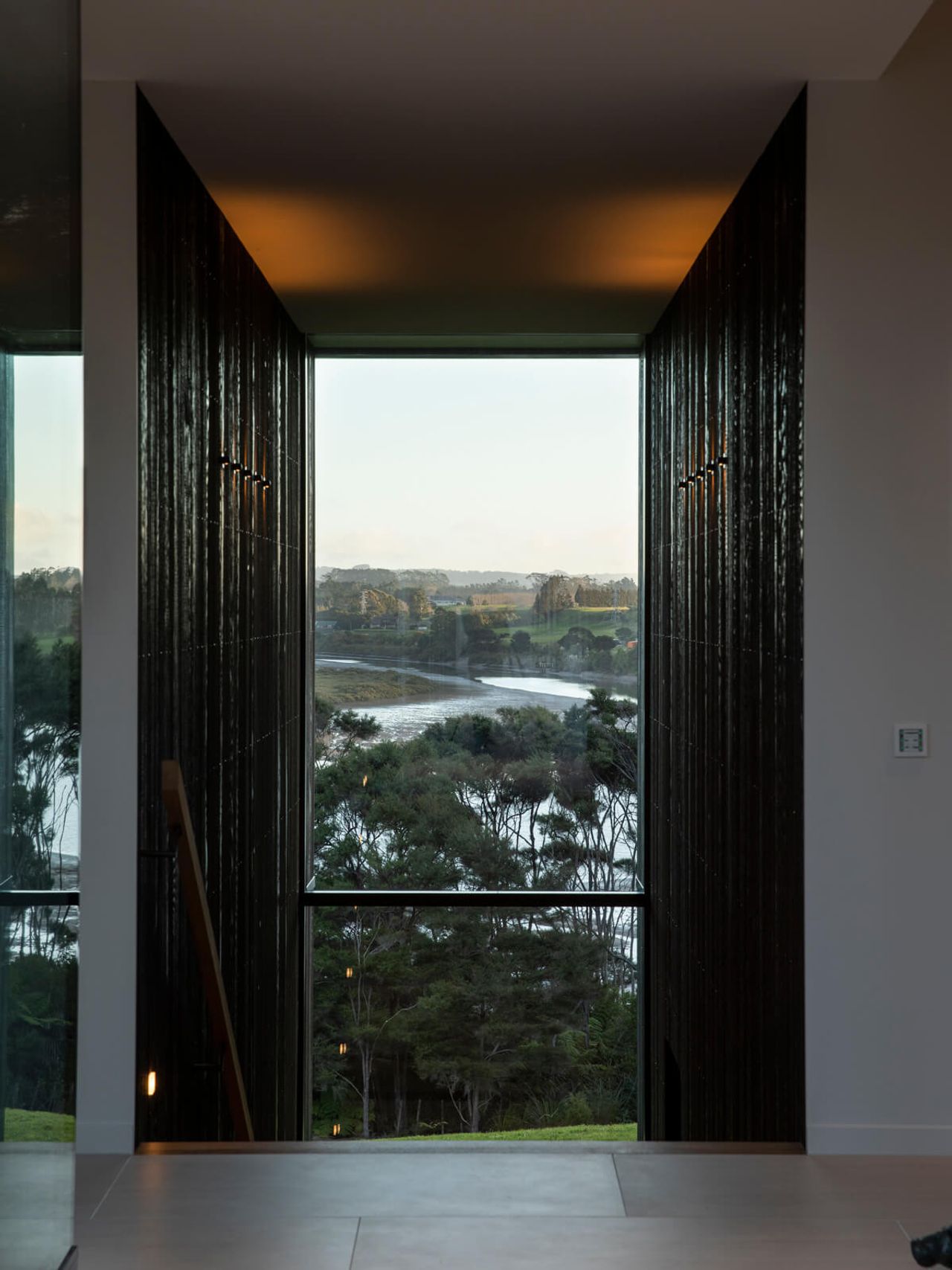 Framed floor-to-ceiling window with breathtaking views of the New Zealand landscape, including lush greenery and a tranquil river, emphasizing modern home architecture and connection to nature.