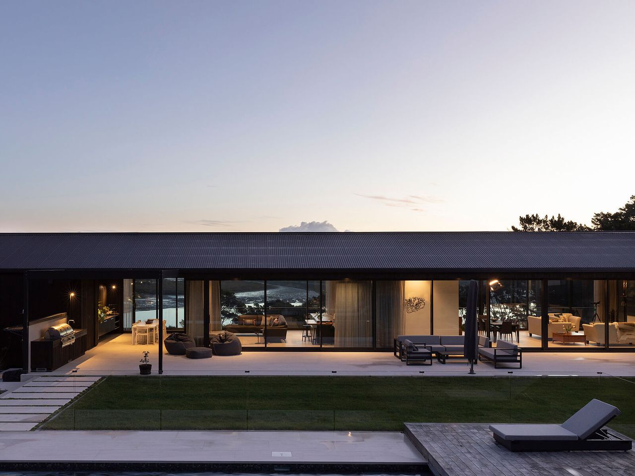 Elegant modern home with open-plan living spaces and floor-to-ceiling glass doors, overlooking a pool and stunning New Zealand river views at dusk.