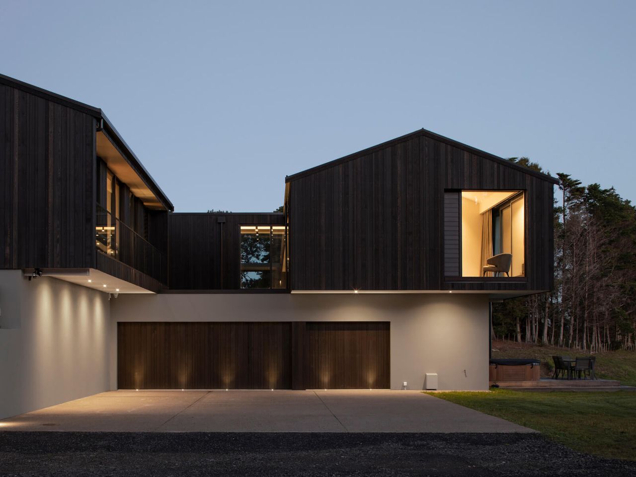 Contemporary two-story home with dark timber cladding and warm interior lighting, featuring a spacious garage and modern architectural design in New Zealand.